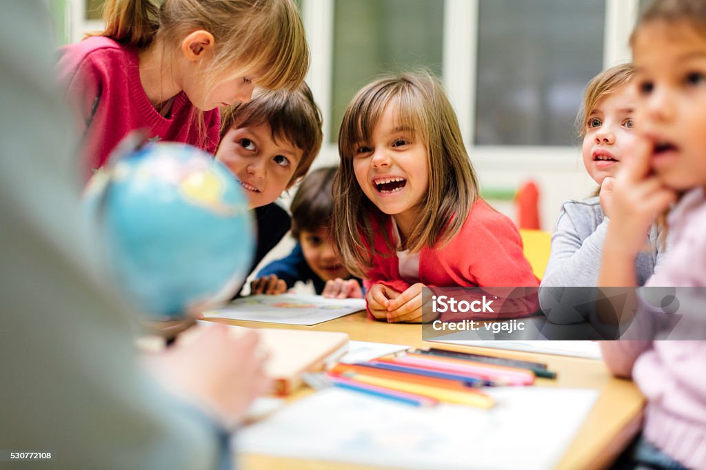 Preschool teacher and children using globe. Preschool teacher teaching her children about geography. Using globe and asking the questions. Children sitting by the table and listen teacher carefully. Models in this shot are part of real kindergarten group and their teacher. Child Stock Photo