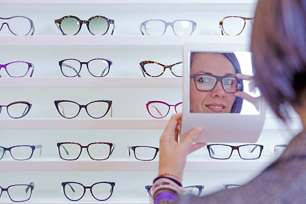 image of a smiling young woman reflected in a small mirror trying on glasses with a glasses exhibitor on background at optical store - focus on left eye