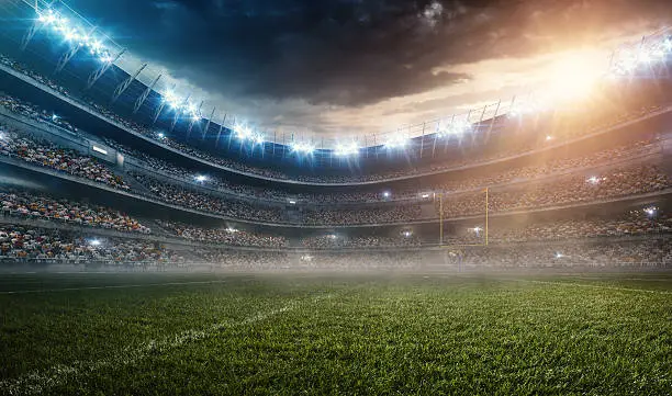 A wide angle panoramic image of a outdoor american football stadium full of spectators under evening sky. The image has depth of field with the focus on the foreground part of the pitch. The view from center of the field.
