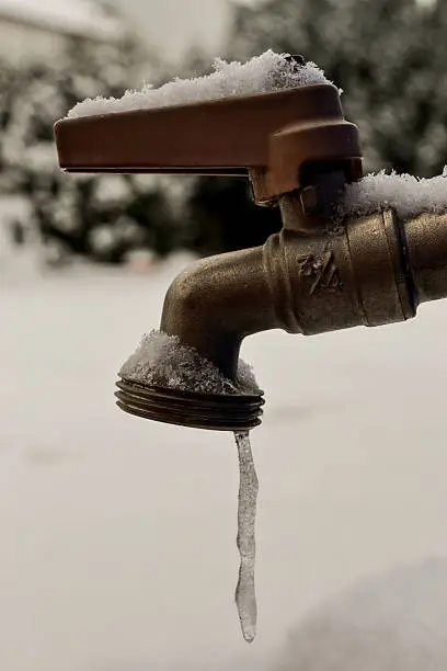 A small amount of water freezes into an icicle as it comes out of a tap, snow covers the ground in this picture. The tap has a snow covered red handle. This picture could be used to symbolize the problems with plumbing in the winter months.