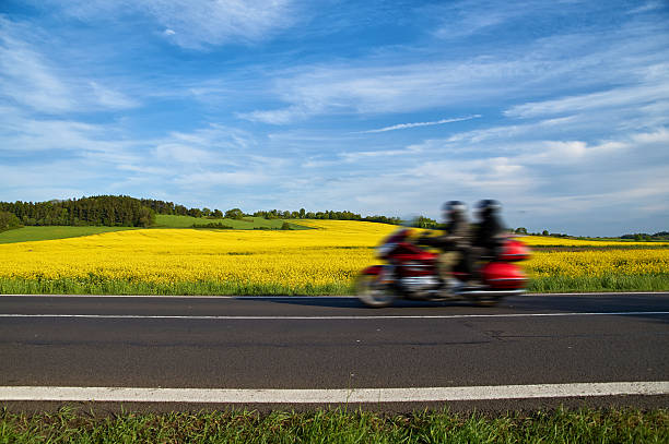 Motion blur red motorcycle speeding on the asphalt road around the yellow flowering rapeseed field. Sunny summer day with blue skies and white clouds.