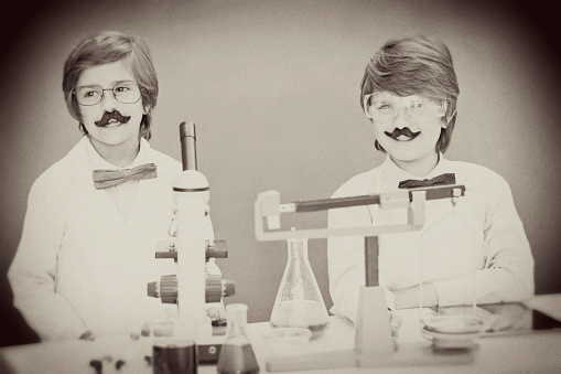 Two excited elementary-age children collaborate on various science experiments.  They are using chemical glassware, chemicals as their experiment is almost ready to test.  Microscope, scale on table.  Imagination, creativity, inspiration for these little chemists!  Chalkboard, classroom setting.  Sepia vingette.