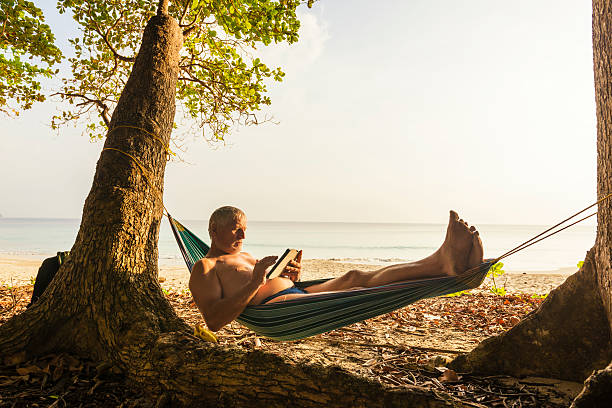 Senior man relaxing in hammock Senior man with digital tablet on hammock between two trees against blue see. Afternoon sunlight.  hammock men lying down digital tablet stock pictures, royalty-free photos & images