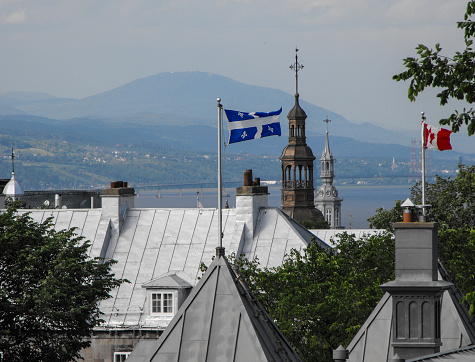 Quebec City rooftop view of church spires, chimneys, flags, the St. Lawrence River, and distant mountain peaks in July.