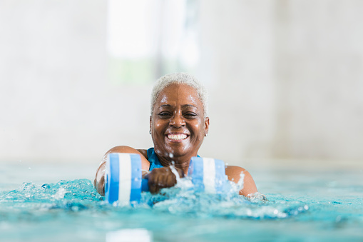 A senior African American woman standing in an indoor swimming pool, doing water aerobics with dumbbells.  Her face, with a big, bright smile, and her shoulders are visible above the water.