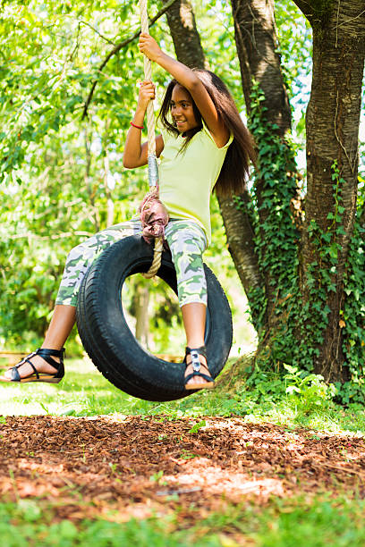 A pair of vibrant pink gymnastic rings hang from the branch of a flowering tree in a backyard garden. The tree is in full spring bloom, surrounded by greenery and a rustic wooden fence. This whimsical and nostalgic scene evokes themes of childhood, outdoor play, nature, and seasonal renewal. Natural light filters through the leaves, casting soft shadows.