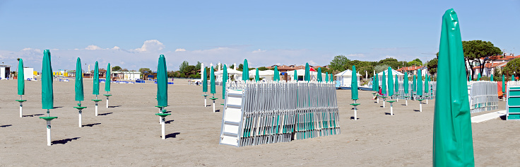 sandy beach with closed sun-umbrellas in green protective covers and closed sun beds, Grado, Italy