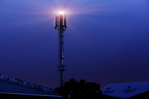 A mobile phone tower with a light on top rises into a deep blue night sky.