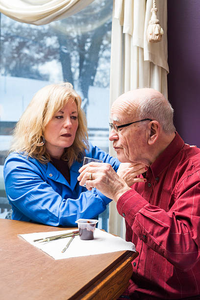 Female speech therapist feels the throat of a senior male patient while swallowing water during a home health therapy session addressing pharyngeal dysphagia