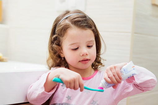 Cute little girl in her pyjamas in bathroom putting a toothpaste on toothbrush