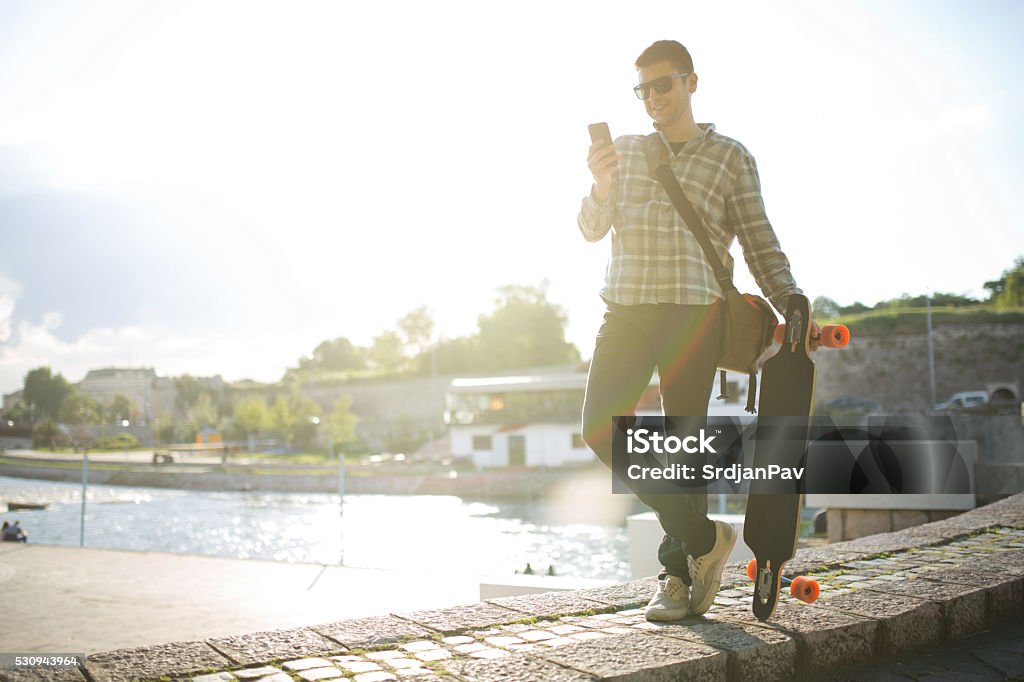 City Boy Young man standing on the quay rampart and looking at his mobile phone, with a longboard in his hand and a bag on his shoulder. Fashionable Stock Photo