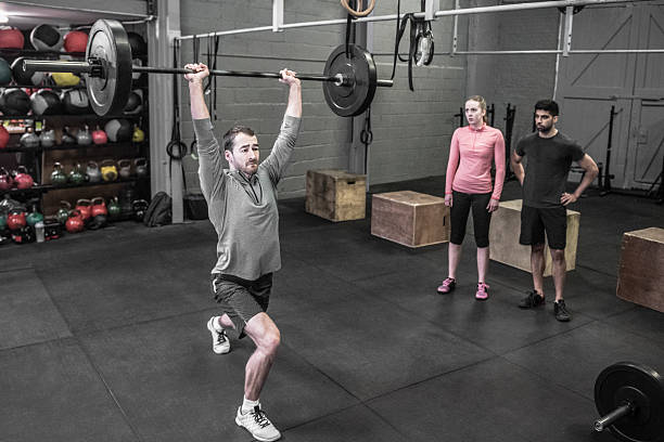 Mid adult man lifting barbell weights in cross training Man exercising in gym. Active male weight lifting with barbell raised above head. Candid portrait of healthy man keeping fit in exercise class. Two friends watching man lift weights. clean and jerk stock pictures, royalty-free photos & images