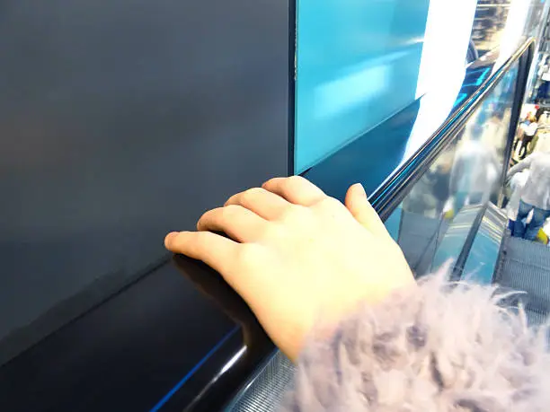 Photo showing a girl's hand on an escalator handrail / rail.  This is a concept image for germs, as research shows that the rail of escalators / moving stairs contains high levels of germs, including the cold and flu virus, and even blood, mucus, E. coli, feces and urine.  Therefore, many shoppers visiting department stores and shopping malls containing escalators avoid touching the handrail or use a hand sanitiser straight afterwards.