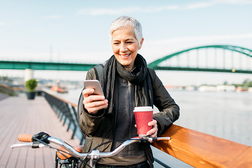Mature businesswoman riding her city bicycle to and from work. She is commuting to work in the morning by riverside. Standing by the fence, typing message on the phone and drinking coffee.