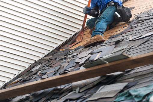 workers remove old asphalt roof shingles with roofing removal tool from suburban home for replacement of new roof tiles.