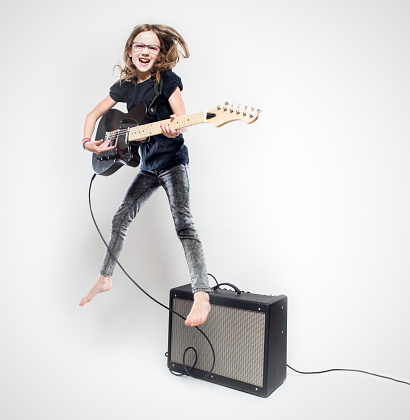 An elementary aged girl plays a black electric guitar on a white studio background.  She laughs with joy as she jumps up in the air while rocking out.  GUITAR IS FULLY RELEASED.