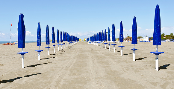 sandy beach with rows of closed sun umbrellas in blue protective covers at Grado, Italy