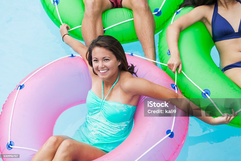 Young woman on float in swimming pool with friend Young woman (18 years) relaxing with friends on innertubes, floating in swimming pool. 18-19 Years Stock Photo