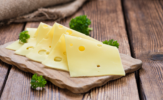 Cheese pieces on a wooden cutting board, on a kitchen table with a large piece of cheese\nin the background, with bread crumbs, large copy space area