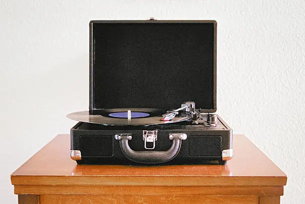 35mm picture of black portable phonograph front view with lid open playing vinyl record sitting on a brown wooden table against a white background.