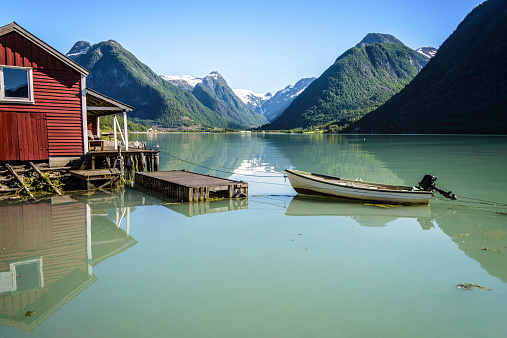 Fjærland, Norway - June 14, 2014: Reflection of a boat, a jetty, a red boathouse and mountains in the tranquil water of Fjærlandsfjord, part of the Sognefjord in the village of Fjærland or Mundal, Sogn og Fjordane, Fjord Norway on June 14, 2014.