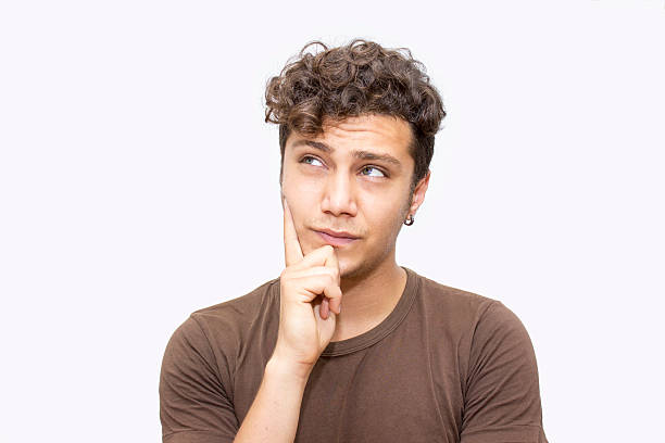 Portrait of thinking young man over white background. Young man standing in front of white background thinking something. One of his hand is on his chin. He has got short,curly brown hair. He is wearing casual clothes. Young man's ethnicity belongs to Turkish ethnicity. Horizontal composition. Photo taken in studio, developed from RAW format.