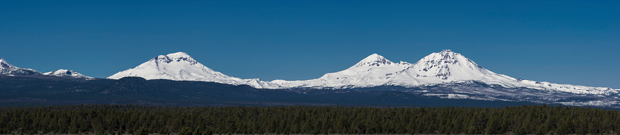 Panorama view of the Three Sisters Mountains, located near the town of Sisters, Oregon. They are located in both Lane and Deschutes counties. The mountains have a nice coating of snow and are framed with trees and blue sky. This is a composite of multiple images.