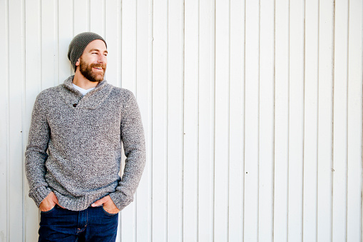 Bearded laughing an leaning against a wall. Wearing a chunky sweater and a hat. Copy space of white wood background.