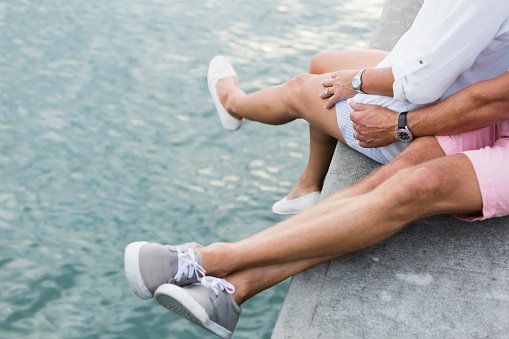 Cropped view of the legs of a mature couple sitting on a dock by the water, legs dangling over the edge.  They are wearing shorts and shoes.
