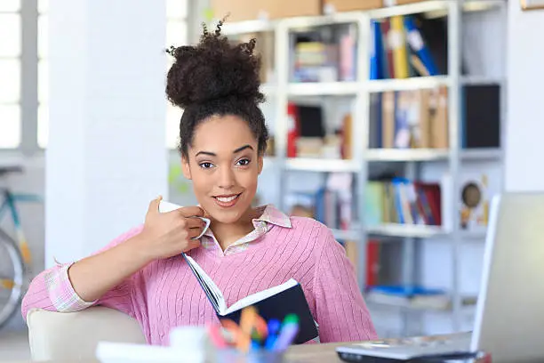 Smiling young woman sitting on her desk at the office. Holding a note book and looking at camera. As background shelves with boxes and folders, bike and tall windows. Office tools,  laptop, landline phone on desk.