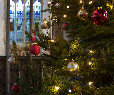 Interior of an English church at Christmas, with a fresh tree hung with red and gold baubles and clear lights. Focus on the baubles, throwing the stained glass window behind the altar into soft focus.
