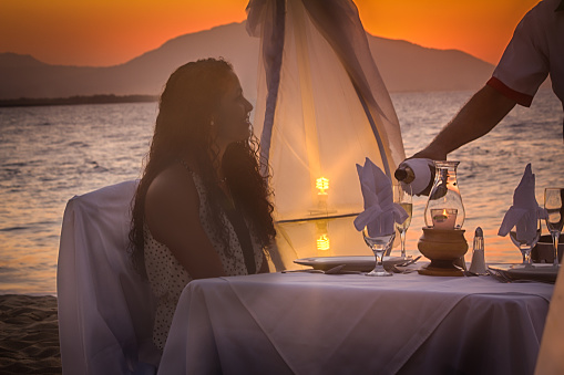A beautiful Caribbean woman from the Dominican Republic enjoys a romantic dinner on the beach at a resort while being served champagne.  