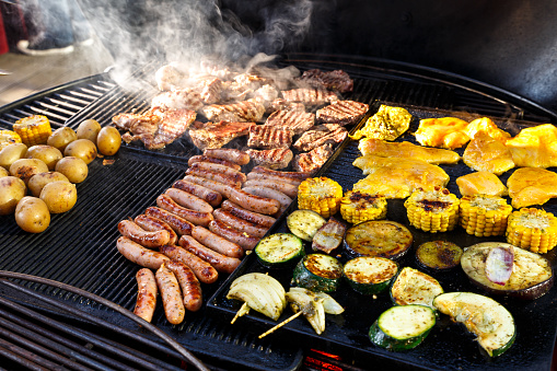 Large hot smokey barbecue with meat, sausages and vegetables being cooked.