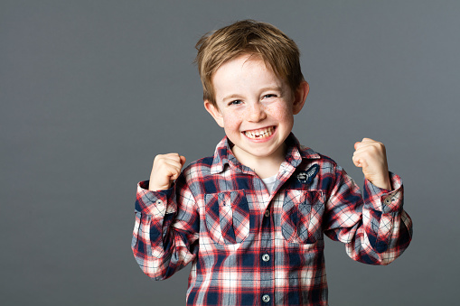winning kid - winning young red hair child with a tooth missing raising his arms for happiness and excitement, grey background studio