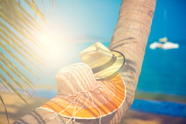 The photo depicts a woman from the back, standing at a beach. She is wearing a yellow straw hat, with a wide brim that shades her head and shoulders from the sun. Her hair is wet, suggesting she has recently been in the water.