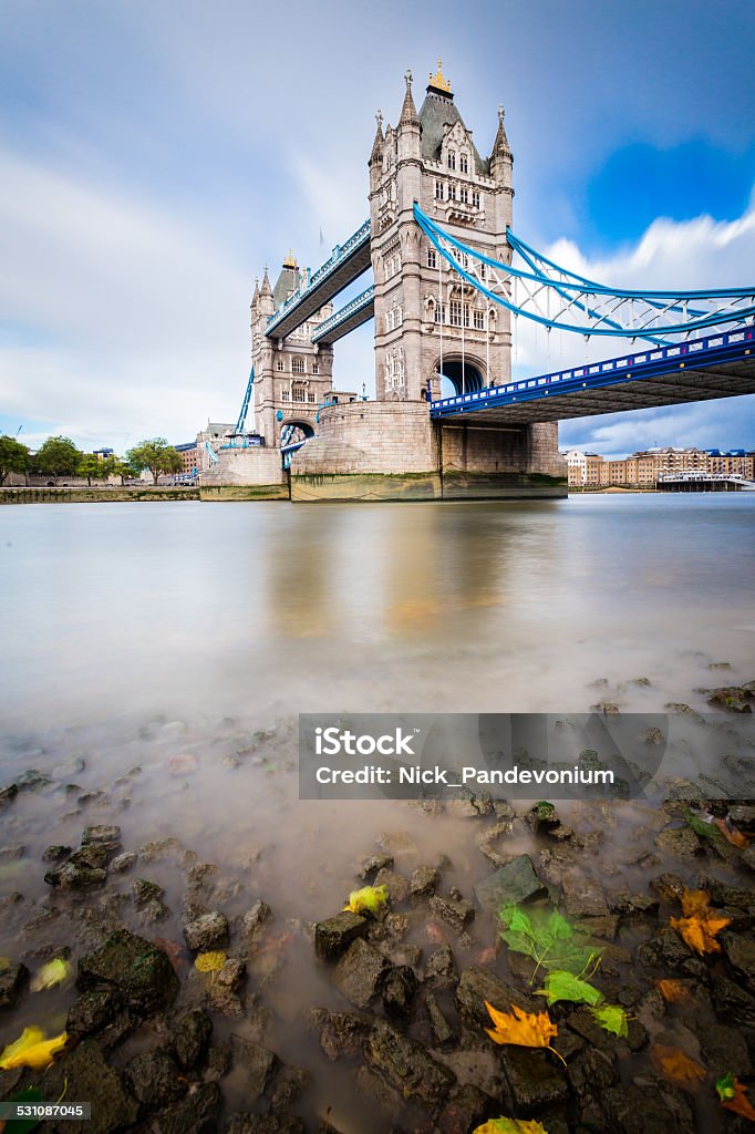 Tower Bridge of London after Saint Jude storm river shore The iconic and famous tourist atraction of Tower Bridge into the city London overlooked from the Southbank of the River Thames at very low angle from the shore with some Autumn coloured leaves and river bed rocks, captured by long exposure technique to emphasize the moving clouds over a peroid of 30 seconds. Shot with Canon EOS 6D, wide angle L lens, ISO 160, long exposure with an ND bigstopper filter, minor vignette added 2015 Stock Photo