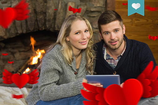 Couple using tablet PC in front of lit fireplace against heart label