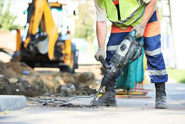 Builder worker with pneumatic hammer drill equipment breaking asphalt at road construction site