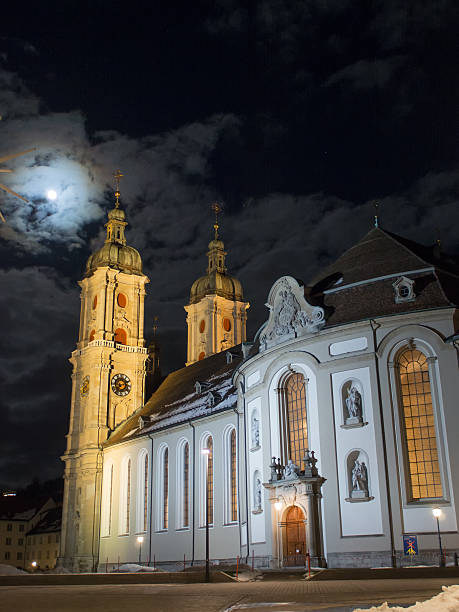 A stunning view of a baroque-style Cathedral of Sankt Gallen with twin towers at sunset, surrounded by snow. A Christmas tree is visible in the foreground.