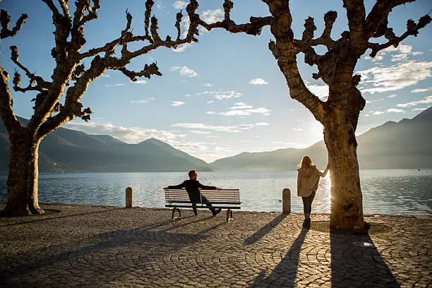 Man sitting on a bench and a woman standing nearby laying on a tree, both looking in a direction of the sunset by the lake. Ascona-Switzerland.