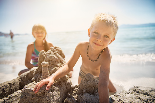 Brother and sister smiling into the camera, having fun building a sandcastle on the beach. They are blonde and wearing swimsuits, backlit bu the summer sun. 