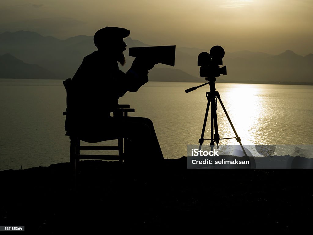 Silhouette Of Adult Man Directing Film In Outdoor Movie Set Silhouette of mid adult film director sitting on director's chair and shouting through megaphone in outdoor movie set.It is sunset time.Orange color is due to down time.There is a camera on tripod in front of him.The model has beard and wearing a flat cap.Sea and mountains are seen on the background.Shot with medium format camera Hasselblad. Director Stock Photo