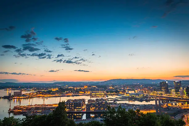 The waterfront landmarks of central Oslo illuminated against the golden glow of summer sunset over the tranquil waters of the Oslofjorden and dark forested hills of southern Norway. ProPhoto RGB profile for maximum color fidelity and gamut.