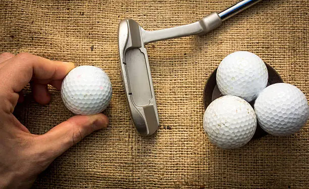 A hand holding a golf ball near a putter and three other golf balls
