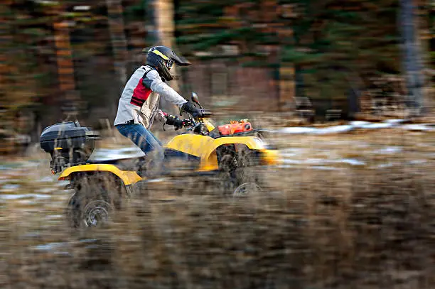 Horizontal motion blurred action shot of a man in helmet and safety goggles riding four-wheeler ATV in snowy autumn forest.