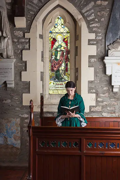 Dark-haired Victorian lady wearing a green dress, standing with bowed head in a church pew reading her prayerbook.