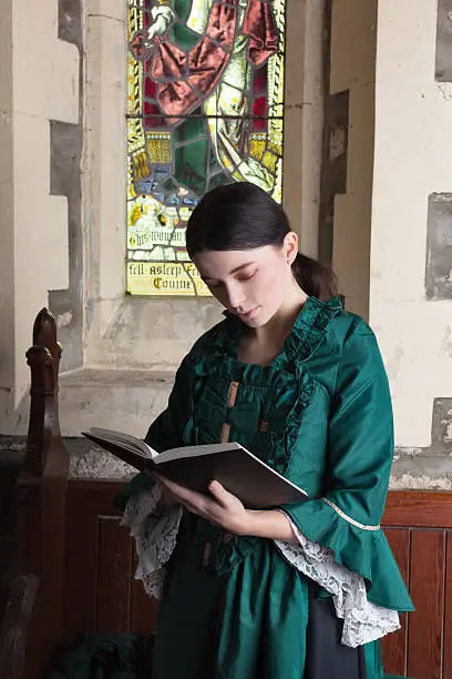 Dark-haired Victorian lady wearing a green dress, standing in a church pew reading from her prayerbook.