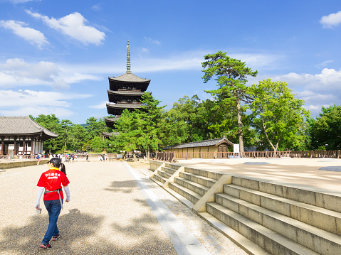 Nara, Japan - August 11, 2014: Back view of young woman in walking alone  in park of an old tourist center in Nara. In background is five store pagoda surrounded with green trees,   some other old buildings and a lot of tourists,  while above them is dynamic cloudy sky.