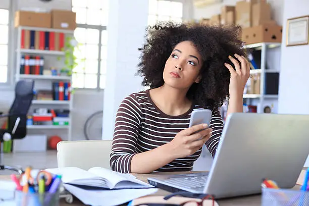 Pensive young woman holding smart phone at workplace. Looking away. As background shelves with boxes and folders, and tall windows with plant, bike and seat. On desk note book, tools, laptop, eyeglasses.