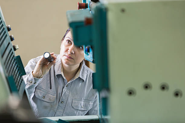 Mid adult woman inspecting equipment in a printing plant with a flashlight.  The printing press is in the foreground.  The worker has a serious expression on her face.